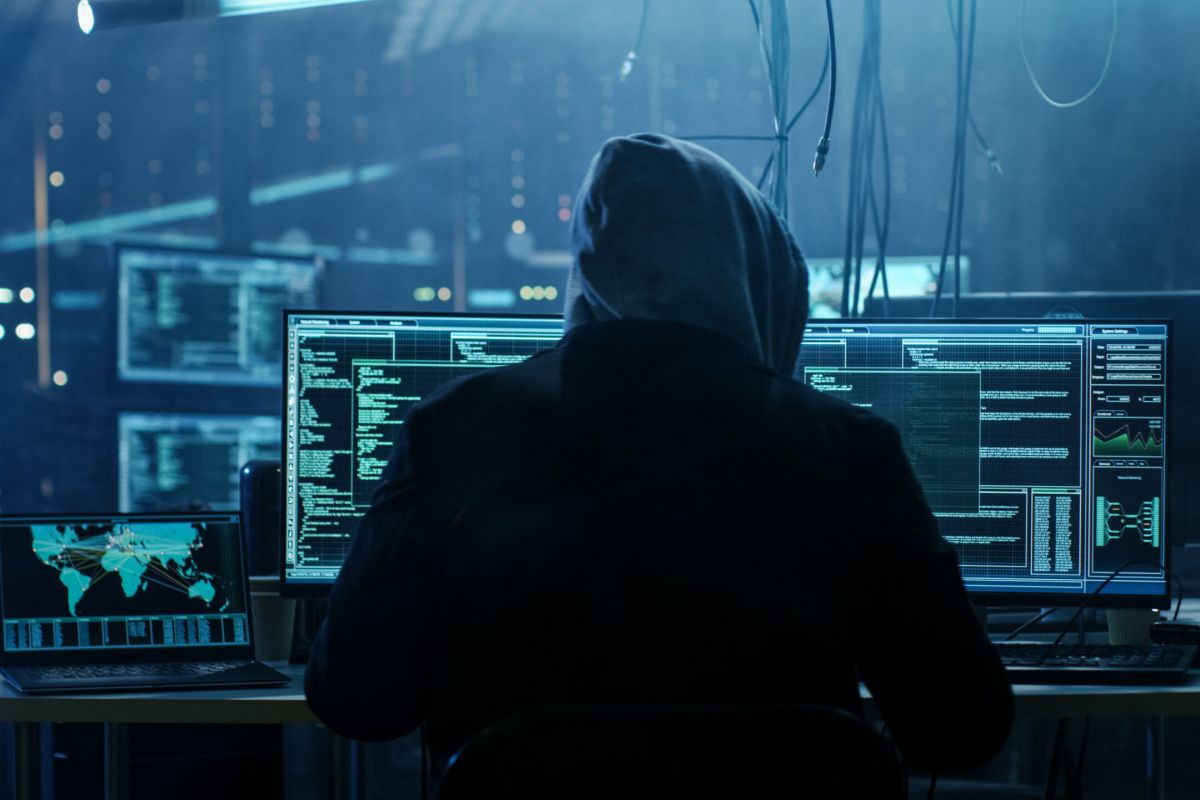 A stock image of a stereotypical black hat hacker at work in a room filled with screens.